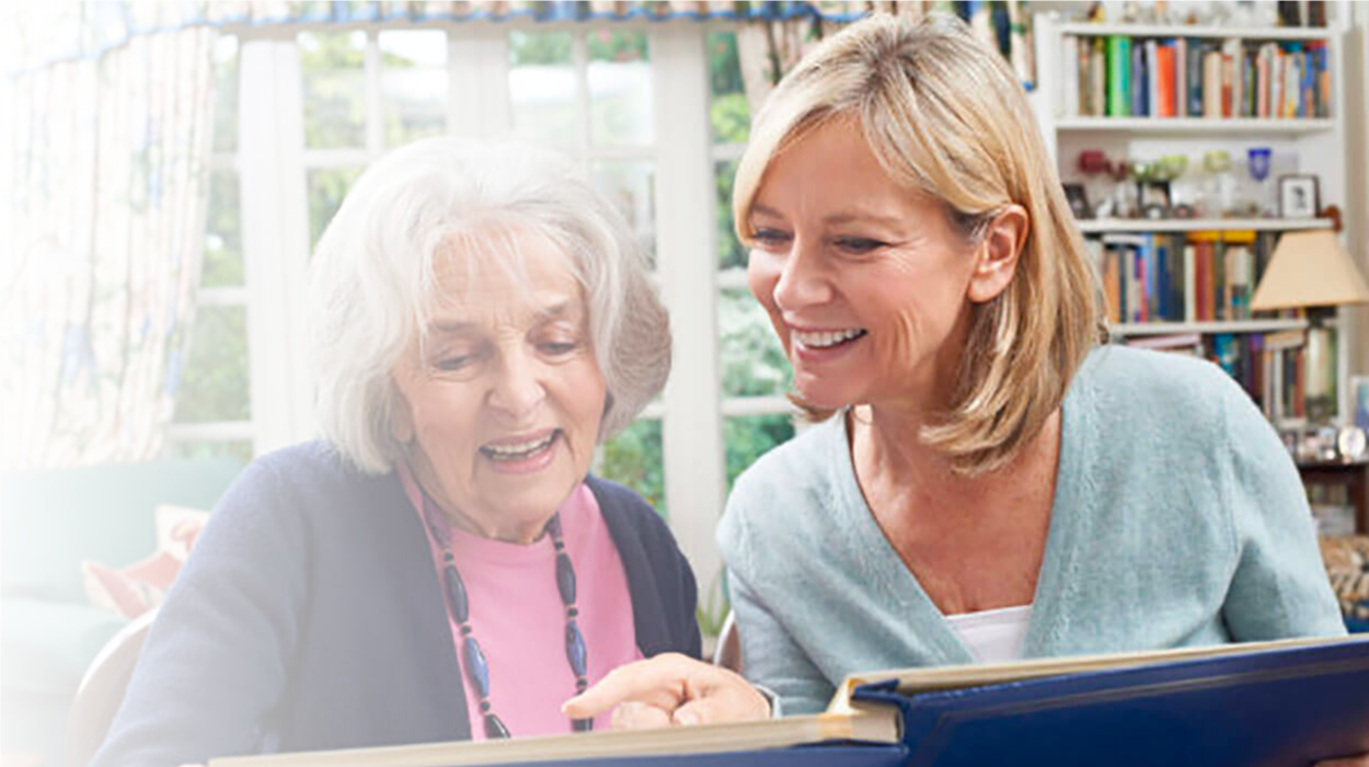 Two women smiling and looking at a large photo album in a cozy living room.