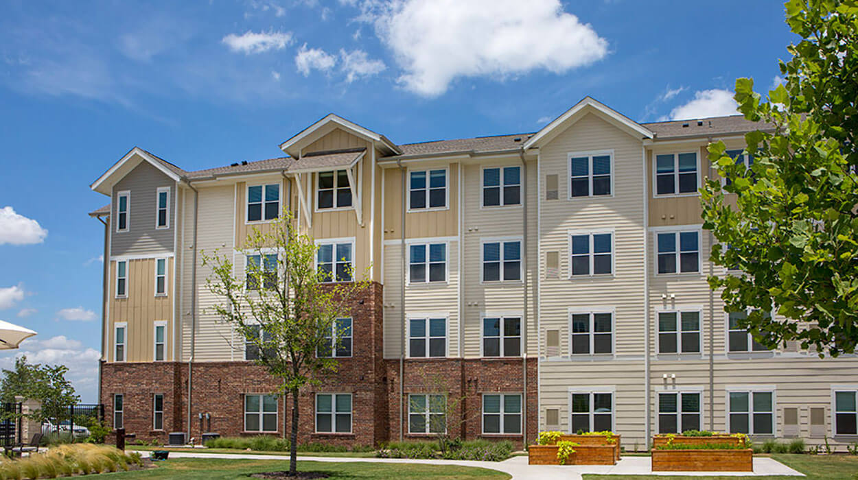 Four-story building with beige siding, manicured lawn, and a few trees on a sunny day.