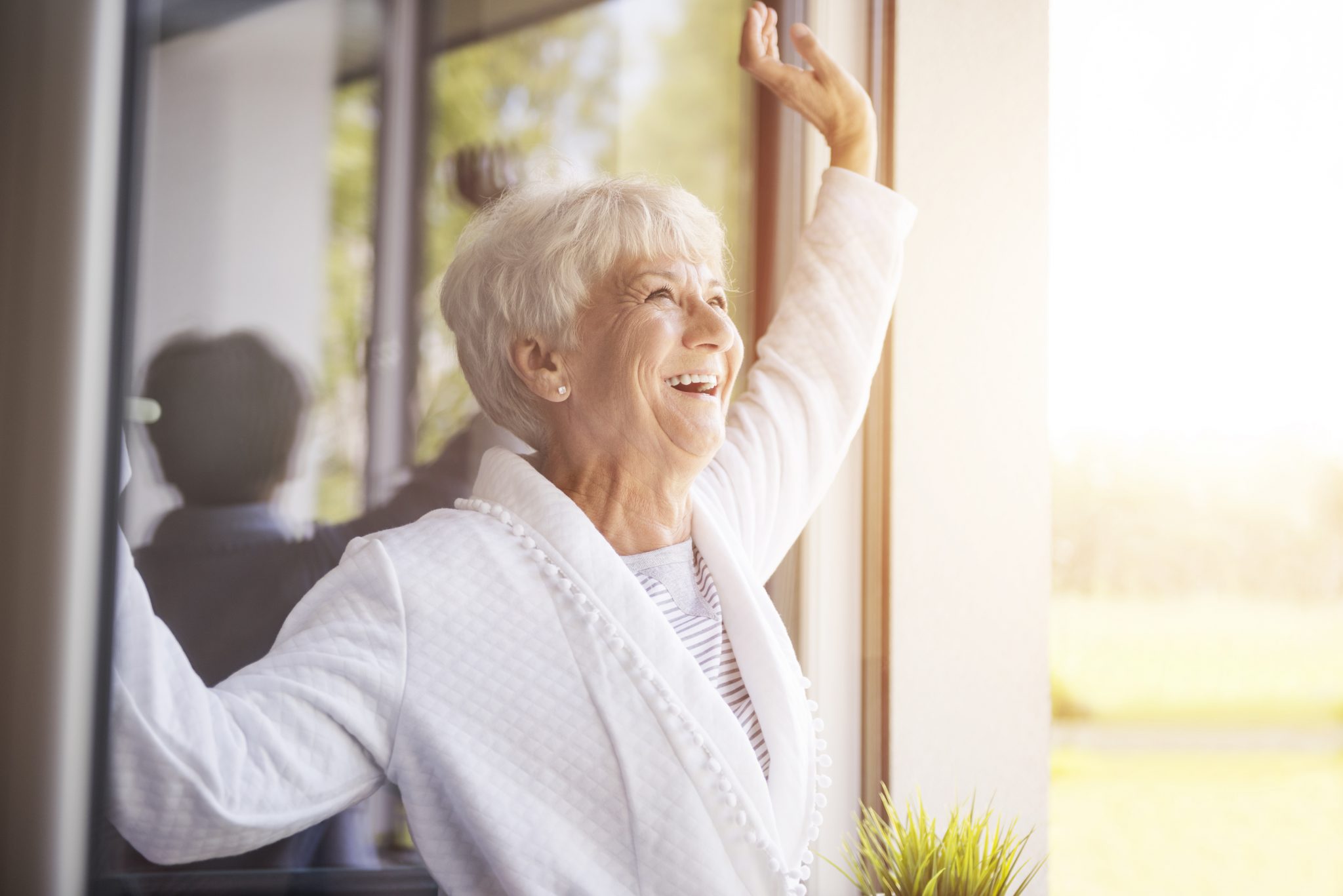 Elderly woman smiling, standing by a sunlit window, dressed in white.