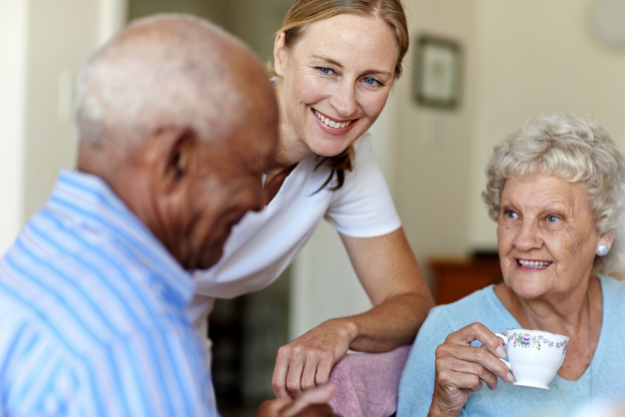 Smiling caregiver interacting with two elderly residents in a cozy living space.