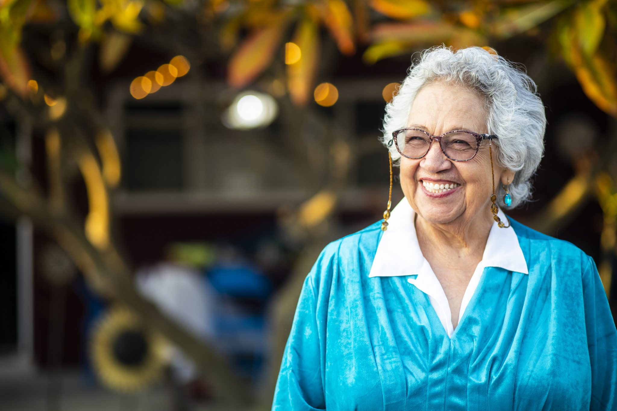 Smiling elderly woman in a blue blouse outdoors with blurred autumn leaves in the background.