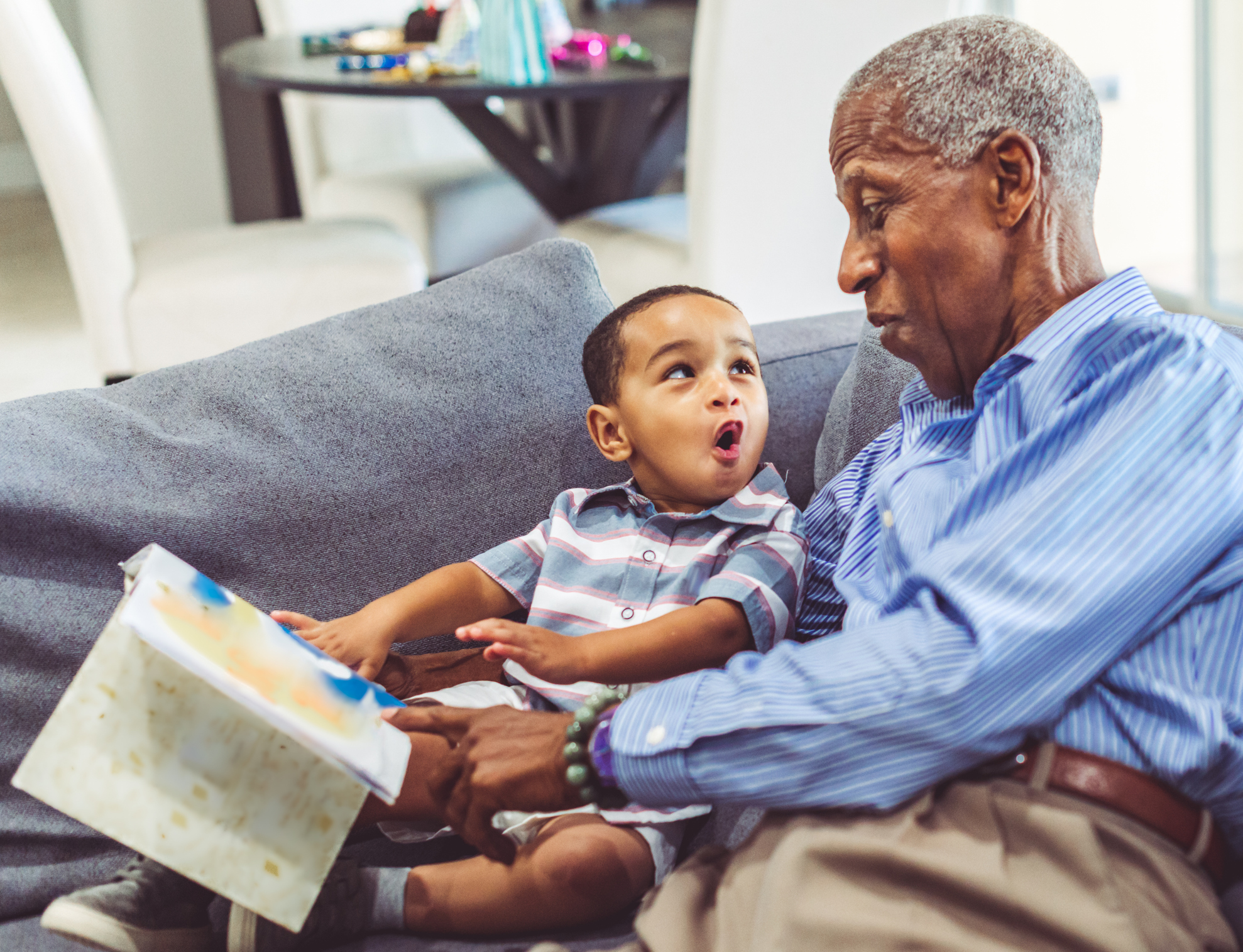 Grandfather reading a book to his surprised grandson on a sofa, sharing a joyful moment.