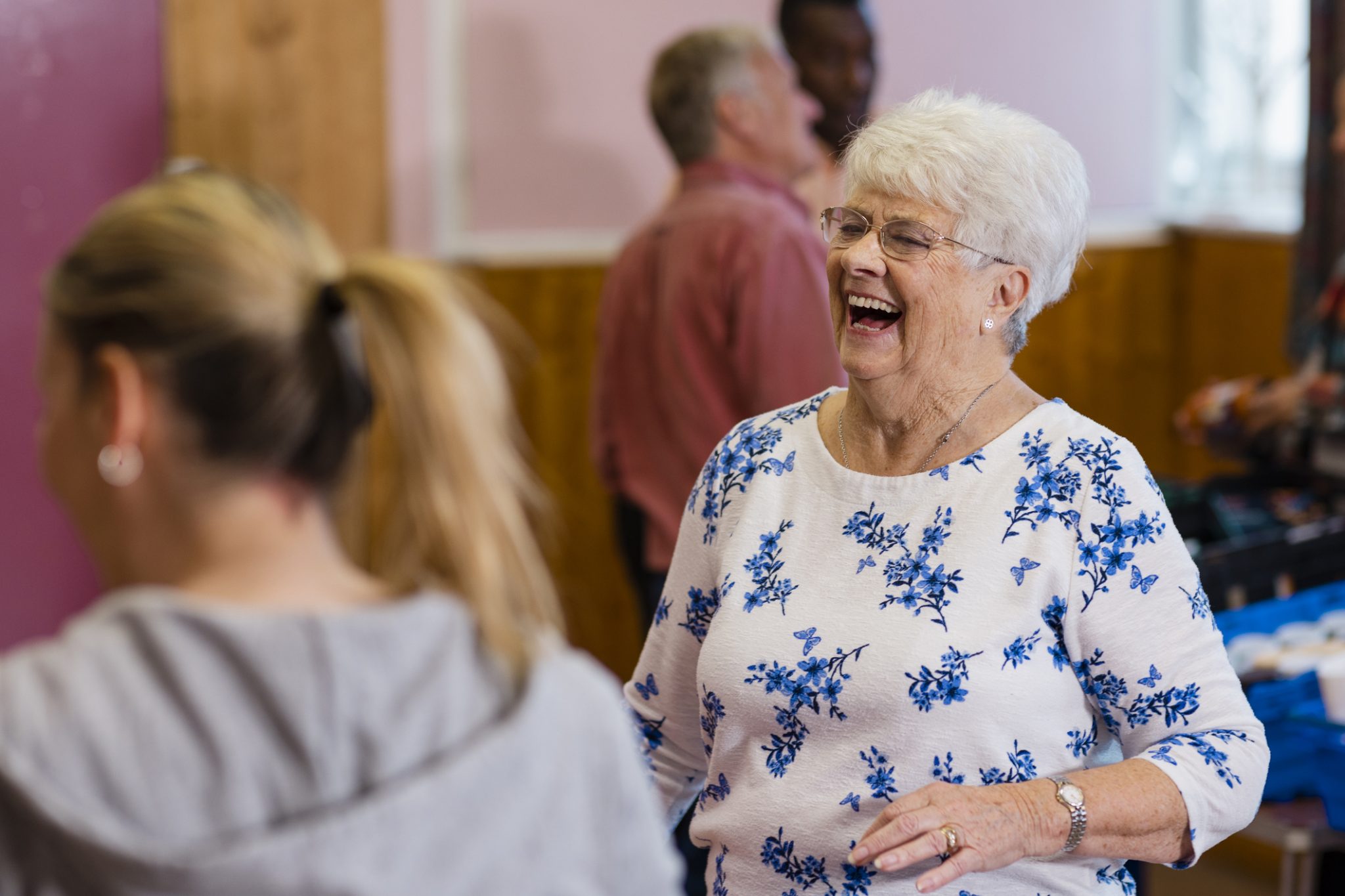 Smiling elderly woman enjoying a lively social gathering with others in the background.