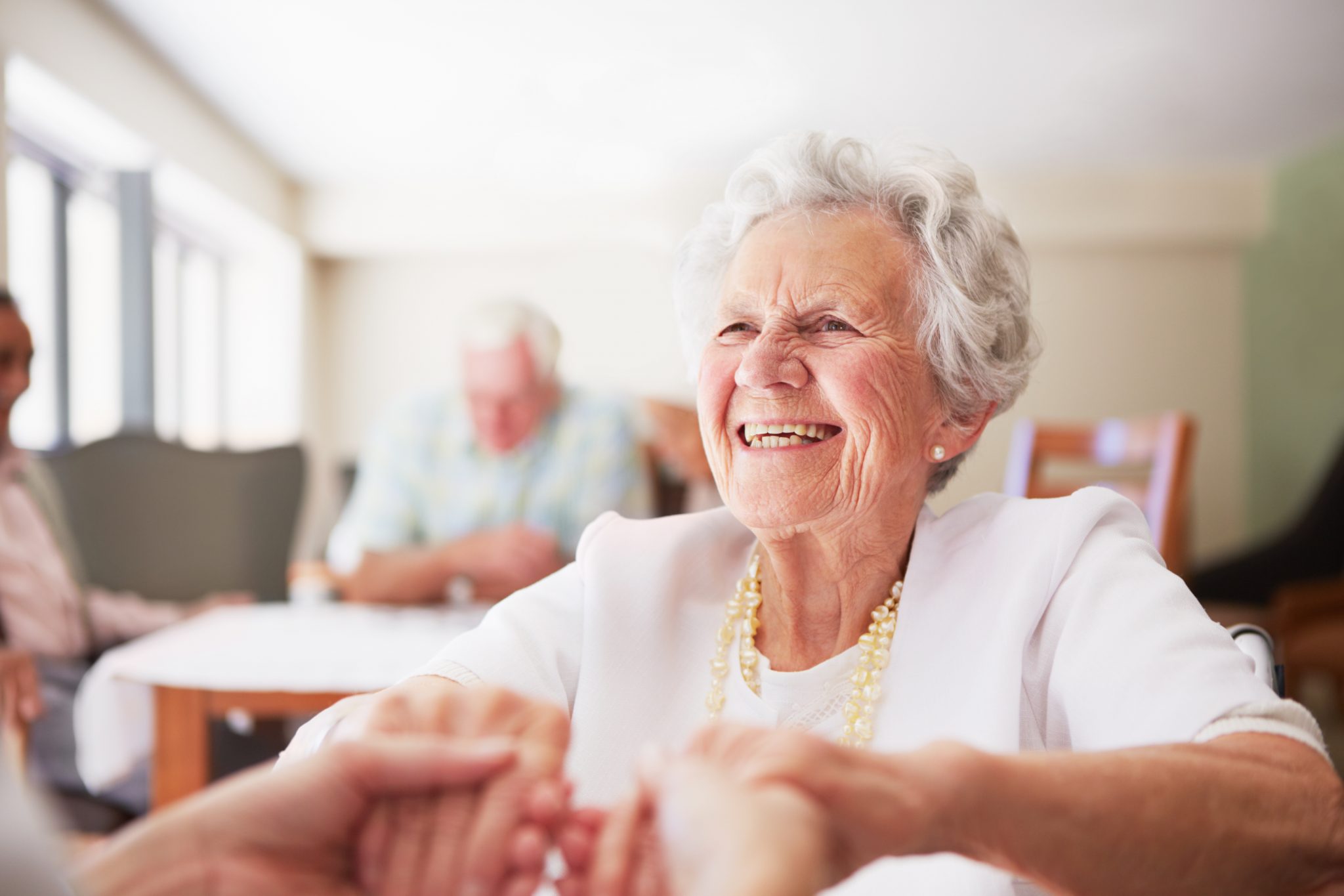 Smiling elderly woman holding hands in a bright communal living space.